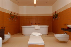 zentralperspektivisch aufgenommenes Badezimmer mit der Wanne im Hintergrund und links und rechts Waschbecken, Toilette und Bidet