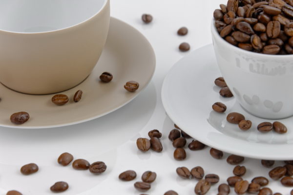 das Bild zeigt zwei angeschnittene Kaffetassen, gefüllt mit Kaffeebohnen, die sich um sie verteilen