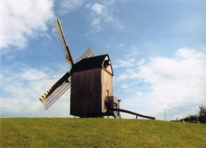 das Bild zeigt eine Bockwindmühle auf einem Hügel vor leicht bewölktem sonnigen Himmel